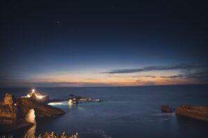 Lit lighthouse on a rocky point under a deep blue dusk sky