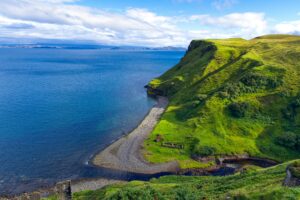 Bright green cliffs dropping to a curved gravel beach beside deep blue water
